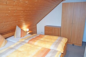 Een slaapkamer met een schuine kap, een bed met een gestreept beddegoed, een bedlampje, een houten garderobe en een klein nachtkastje.