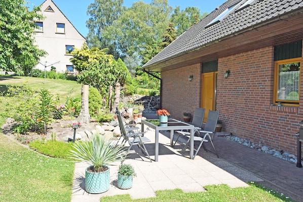 Et murstenshus med tegltag, en have med plantekasser og en terrasse med udendørs møbler.