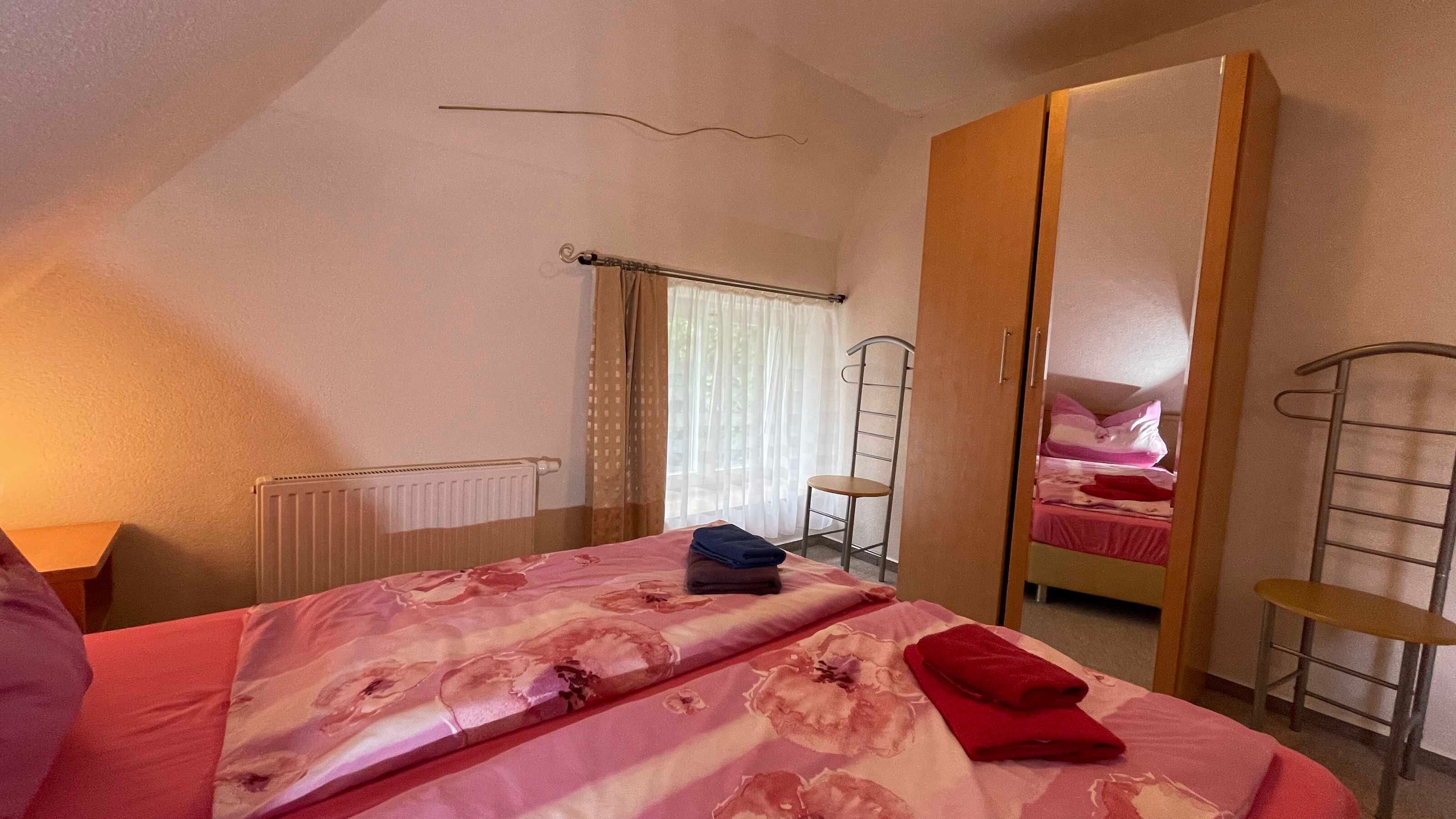 Een slaapkamer met een bed, een kledingkast, een radiator, een raam met gordijnen en een stoel.