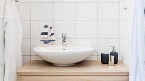 A modern bathroom with a large white sink, a small sailboat decoration, and neatly folded towels on a wooden shelf. - 2-room app. No. 2-3, 34 m², 1st floor, balcony - Gästehaus Schäfersruh (Scharbeutz)