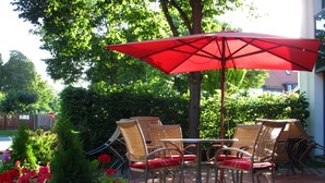 Un patio al aire libre con sillas de mimbre y un paraguas rojo, rodeado de plantas y flores en macetas.