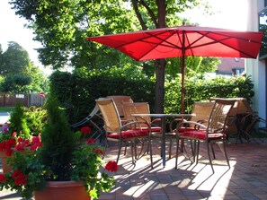 Patio esterno con sedie in vimini e un ombrello rosso, circondato da vasi di piante e fiori.