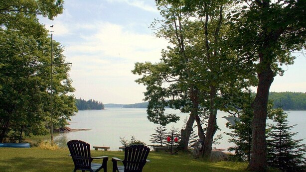 A lakeside view with two Adirondack chairs and a picnic table, surrounded by trees and a clear sky. - Pat's Place - Peacefull & charming cottage (Deer Isle)