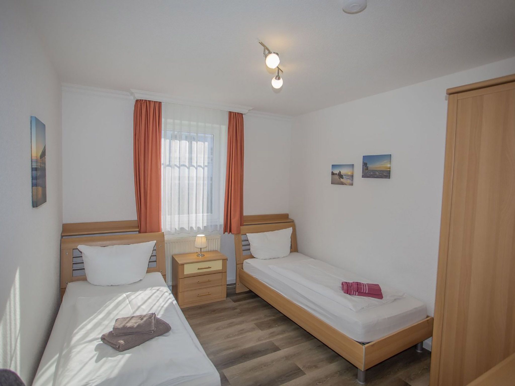 Una habitación con dos camas individuales, un armario de madera, una mesita de noche con lámpara y una ventana con cortinas.