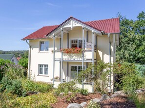 A two-story house with a red-tiled roof, a balcony with flowers, and a garden with various plants.
