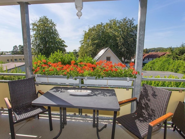 A covered outdoor seating area with two chairs and a table, overlooking a garden with red flowers and a building in the background.