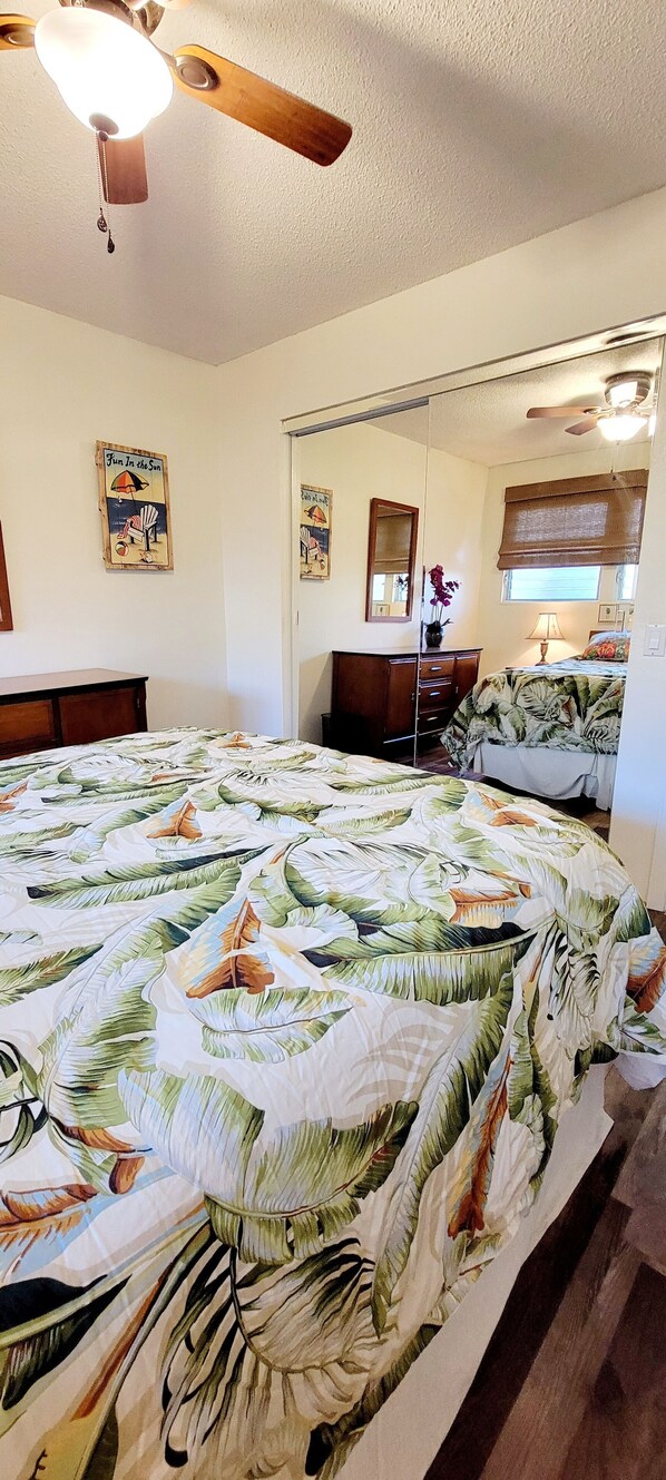 A bedroom with a large bed, a ceiling fan, a dresser, a mirror, and a window with a view. - POPULAR ON OCEAN COMPLEX - GREAT AMENITIES, DISCOUNTS AND LOCATION!!! (Kailua-Kona)