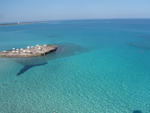 An aerial view of a small island with clear turquoise waters, a sandy beach, and several white umbrellas.