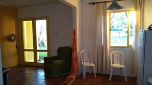 A room with a tiled floor, a green armchair, two white plastic chairs, a hanging light fixture, a window with curtains, and a door with blinds. - Casa del Sol de Itapiruba (Itapiruba)