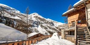 A wooden chalet with a stone staircase and a wooden deck, surrounded by snow-covered mountains.