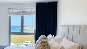 Habitación de hotel con una cama grande, vista al mar y un balcón con coches aparcados fuera.