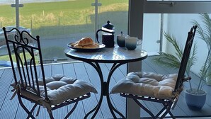 Un balcón con una mesa lista para el desayuno, con vistas a un paisaje costero donde se divisa un castillo a lo lejos.