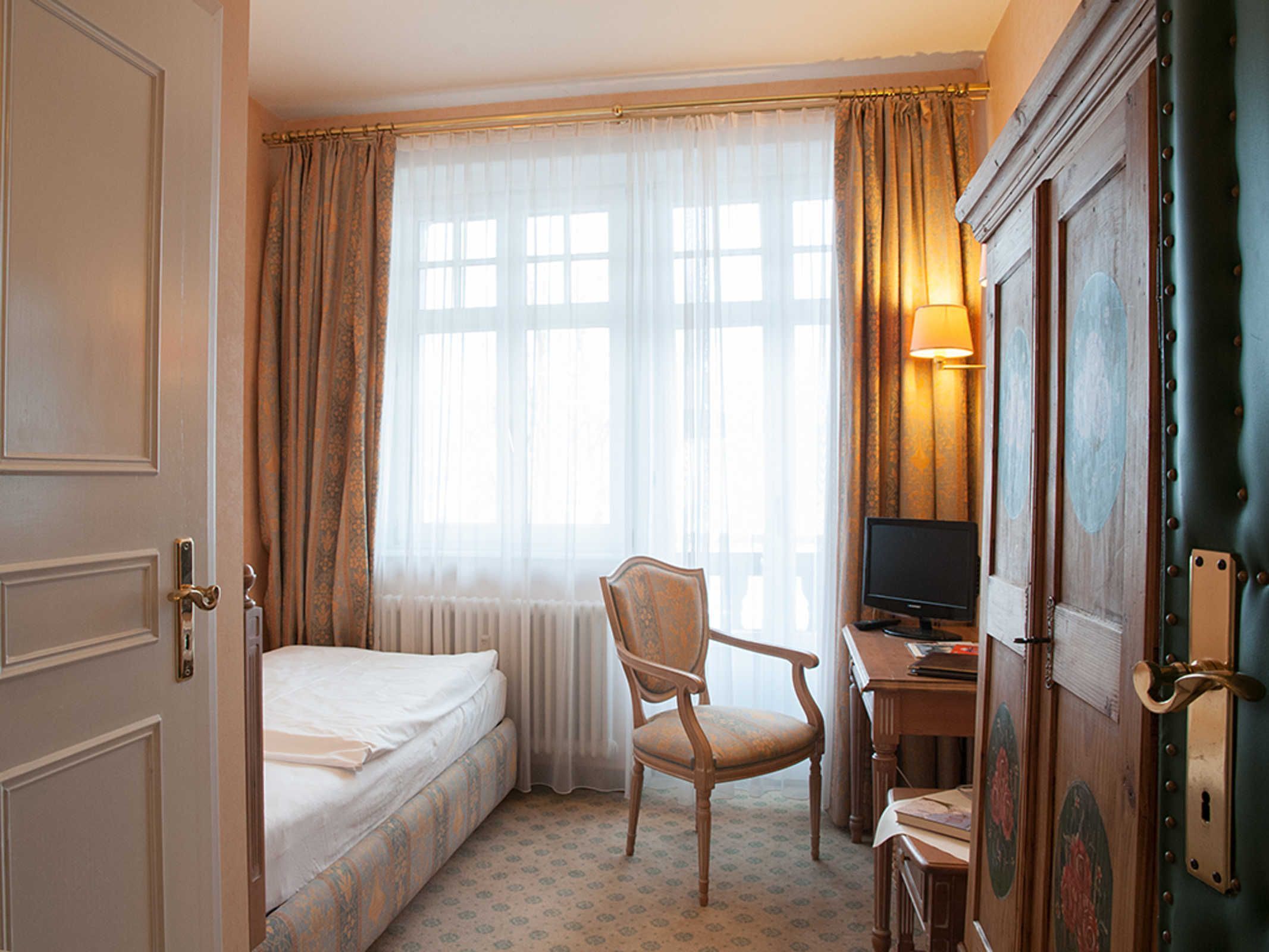 A hotel room with a bed, a chair, a desk with a computer, and a window with curtains.