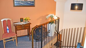 A room with a wooden chair, a small table with a vase of flowers, a framed painting of a ship on the sea, and a staircase with striped wallpaper.