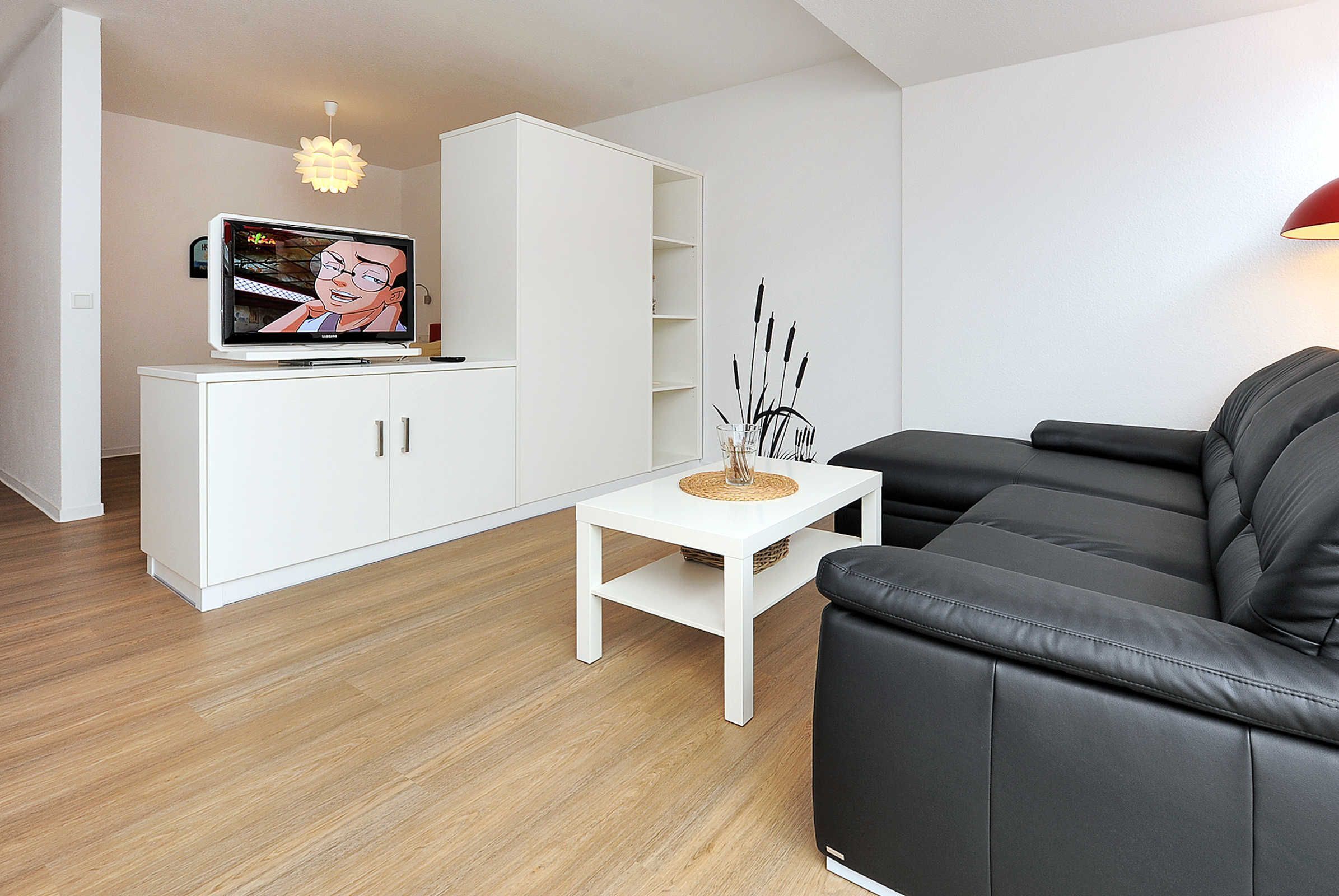 A modern living room with a flat-screen TV, a white cabinet, a black leather sofa, and a small white table.