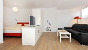 Ein moderner, kompakter Wohnraum mit Bett, Fernseher, einem weißen Schrank, einer schwarzen Couch und einem kleinen Tisch.