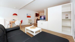 Ein modernes Wohnzimmer mit einer schwarzen Couch, einem weißen Couchtisch und einem Fernseher. Es gibt einen Essbereich mit Tisch und Stühlen, und im Hintergrund eine Küche.