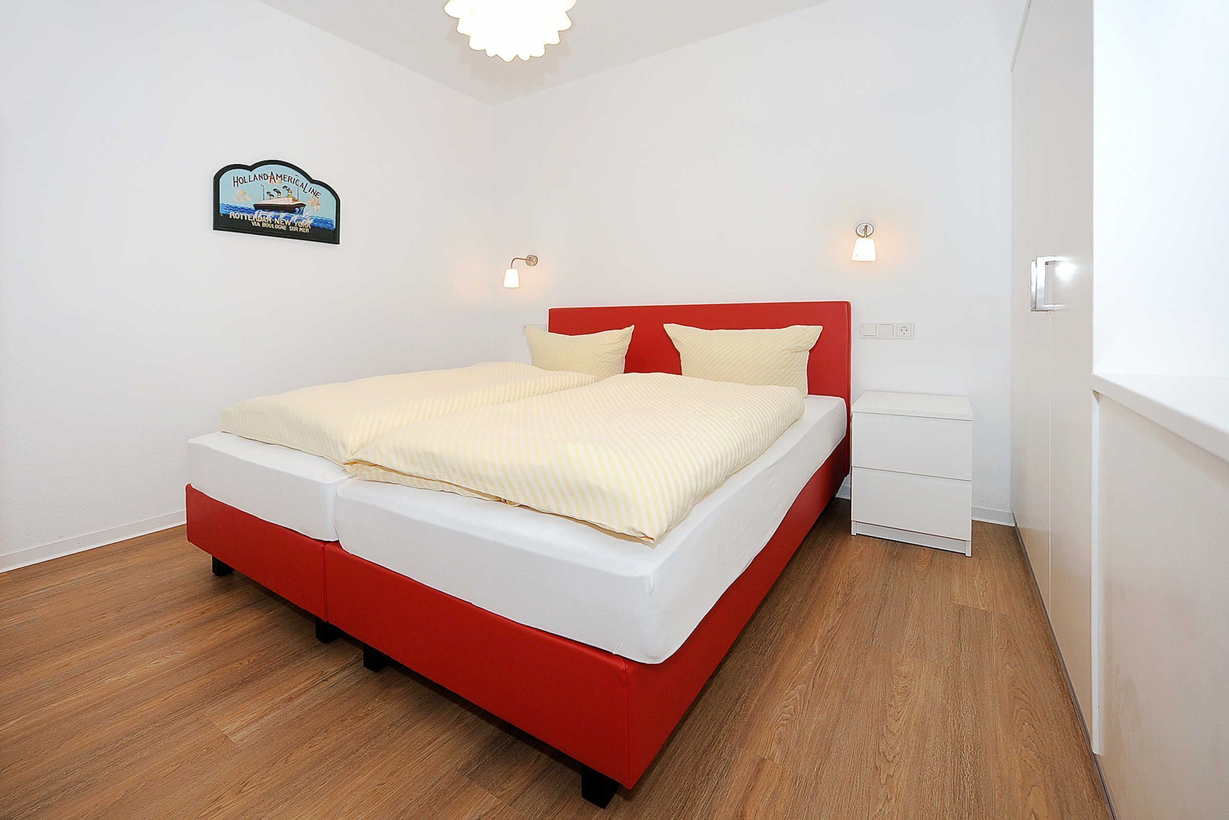 Een bed met een rode hoofdbord en witte beddengoed, een klein nachtkastje en een wandlamp.