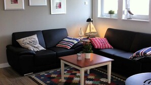 En stue med en sort sofa, et træbord og et farverigt tæppe. Tre billeder hænger på væggen, og et vindue viser en fugl udenfor.