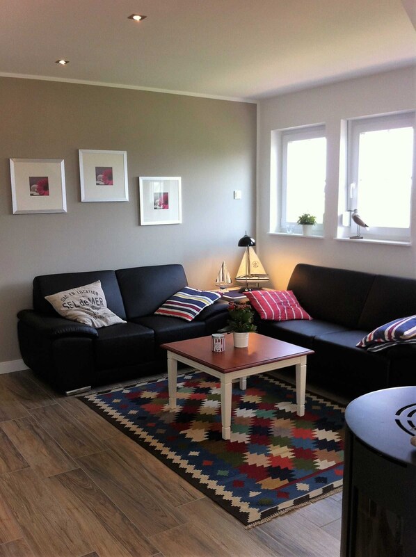 Un salon avec un canapé noir, une table basse en bois et un tapis coloré. Trois tableaux encadrés ornent le mur, et une fenêtre donne sur l’extérieur, où l’on peut apercevoir un oiseau.