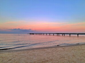 Ein Strand mit einem Steg, der sich in ruhiges Wasser erstreckt, unter einem wolkenlosen Himmel.