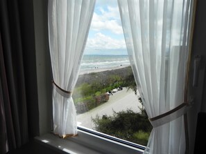 Ein Fenster mit transparenten weißen Vorhängen, Blick auf einen Strand mit Spaziergängern und einem klaren Himmel.