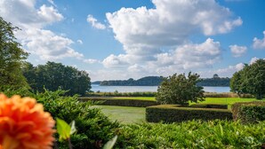Ein angelegter Garten mit Seeblick, ordentlich gestutzten Hecken und einer markanten orangefarbenen Blume im Vordergrund.