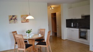 A dining area with a wooden table and chairs, a hanging light fixture, and a kitchen with white cabinets and a black countertop. - Vacation apartment - Kur & Golf Ferienwohnungen Wattenbach (Bad Windsheim)