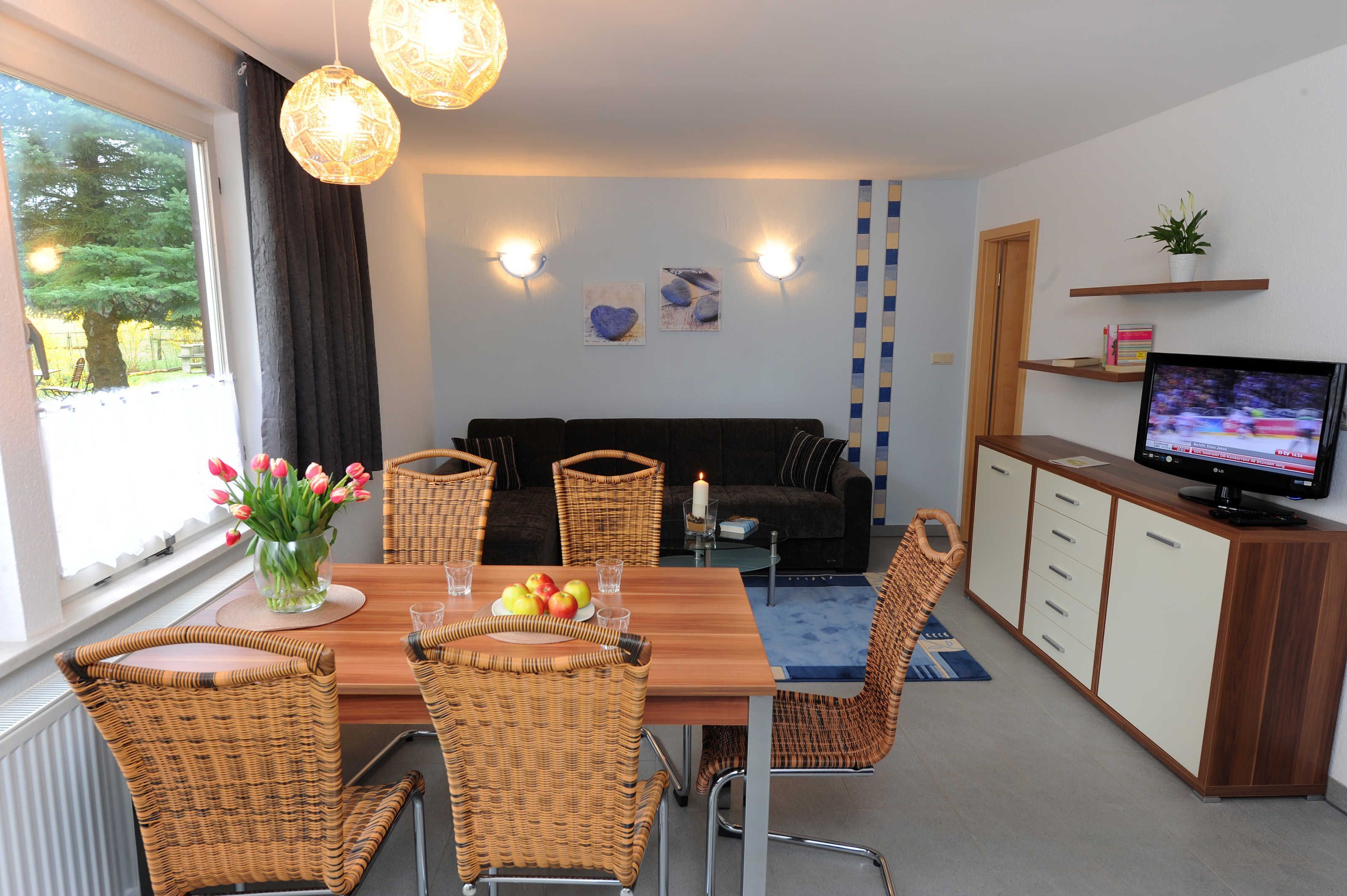 Un salón moderno con una mesa de comedor de madera, sillas de mimbre, un sofá negro, un televisor y una ventana con vistas a árboles.