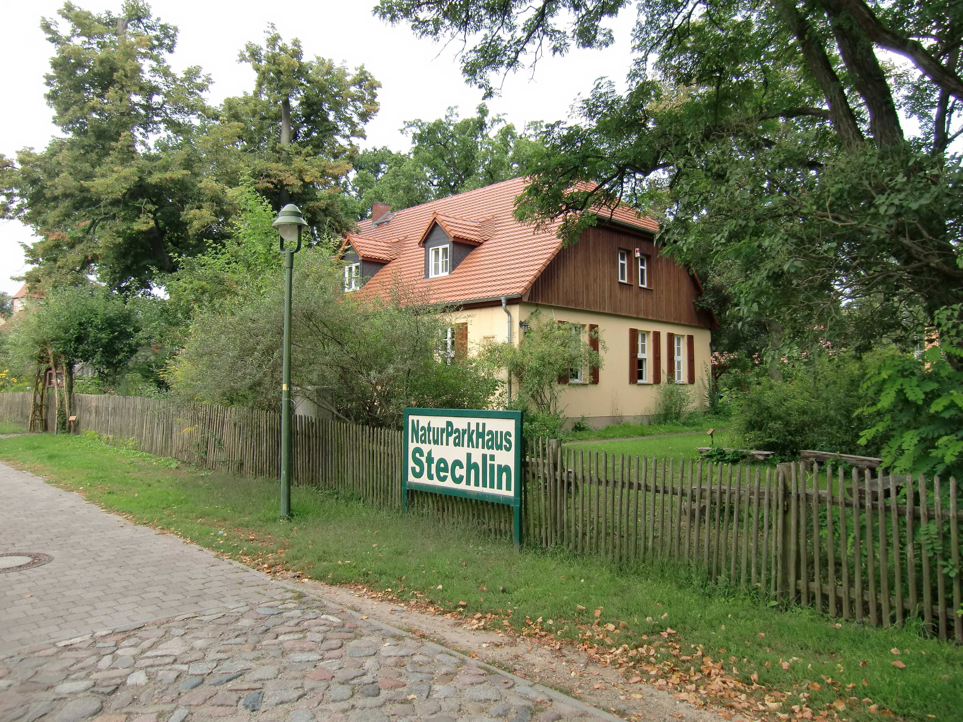Een huis met een bord waarop staat 'NaturParkHaus Stechlin', omringd door groen en een houten hek.