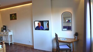 A room with a television mounted on the wall, a small table with a chair, a coffee machine, and a potted plant.