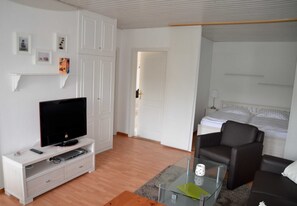 Un salon moderne avec un téléviseur à écran plat posé sur un meuble blanc, une table basse en verre, un canapé en cuir marron et un lit avec du linge de maison blanc dans la chambre adjacente.