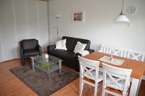 Un salon avec un canapé noir, une table basse en verre, une table à manger blanche avec des chaises, un lampadaire et une horloge murale.
