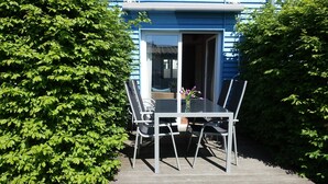 Een eethoek buiten met een tafel en stoelen, een blauw gebouw op de achtergrond en een grote struik aan de linkerkant.