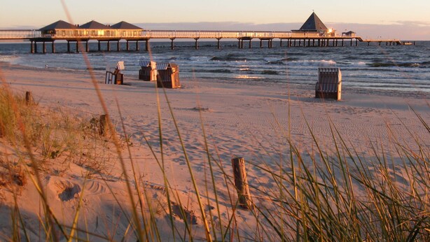 Een strand met een pier die zich uitstrekt in het water, met een gebouw met puntige dak en verschillende kleinere constructies.