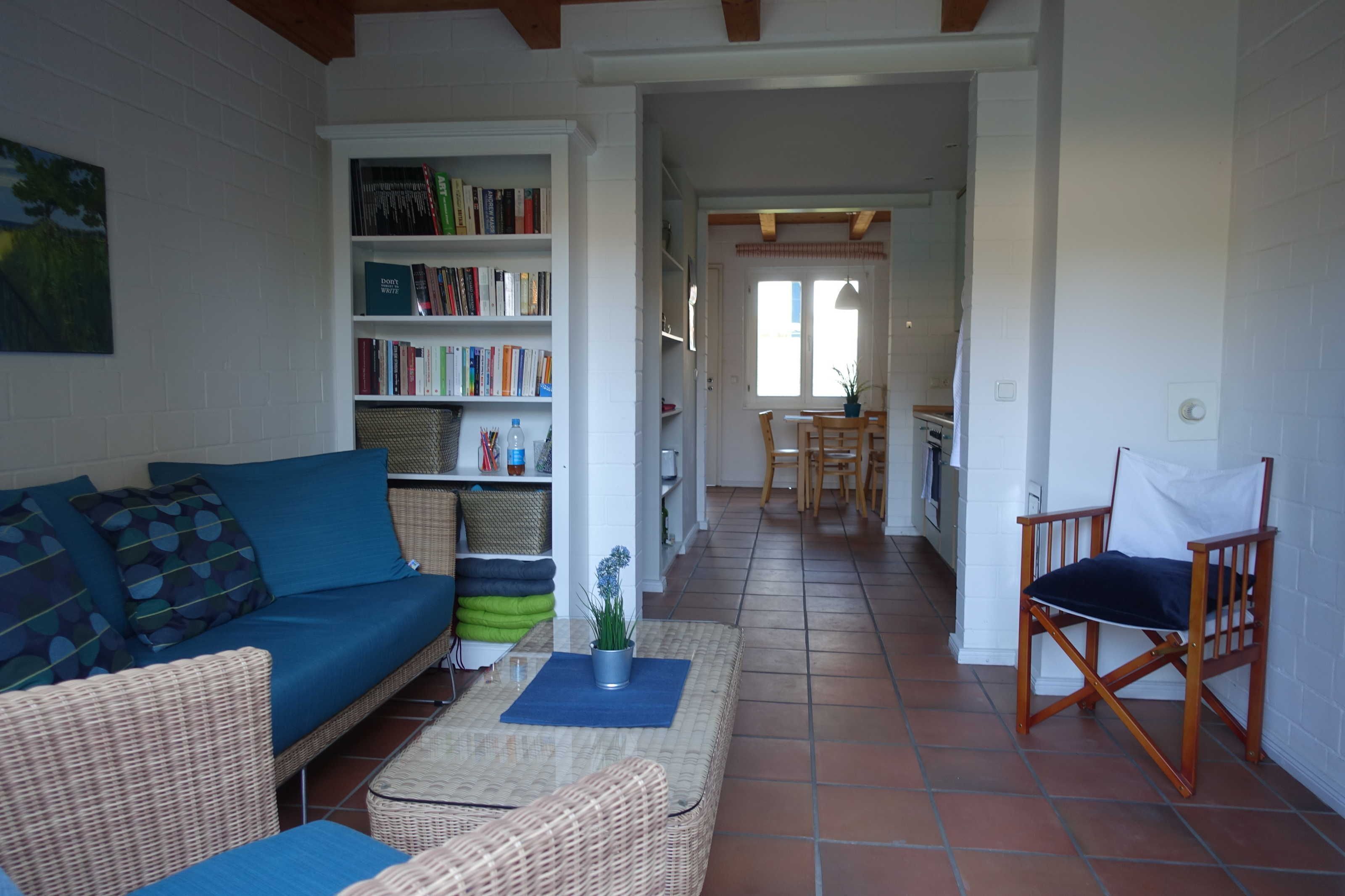 Una sala de estar acogedora con un sofá azul, sillones de mimbre, una estantería y una zona de comedor con una mesa y sillas.