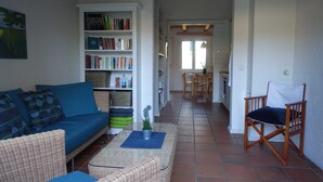 Een gezellige woonkamer met een blauwe bank, rieten stoelen, een boekenkast en een eethoek met een tafel en stoelen.