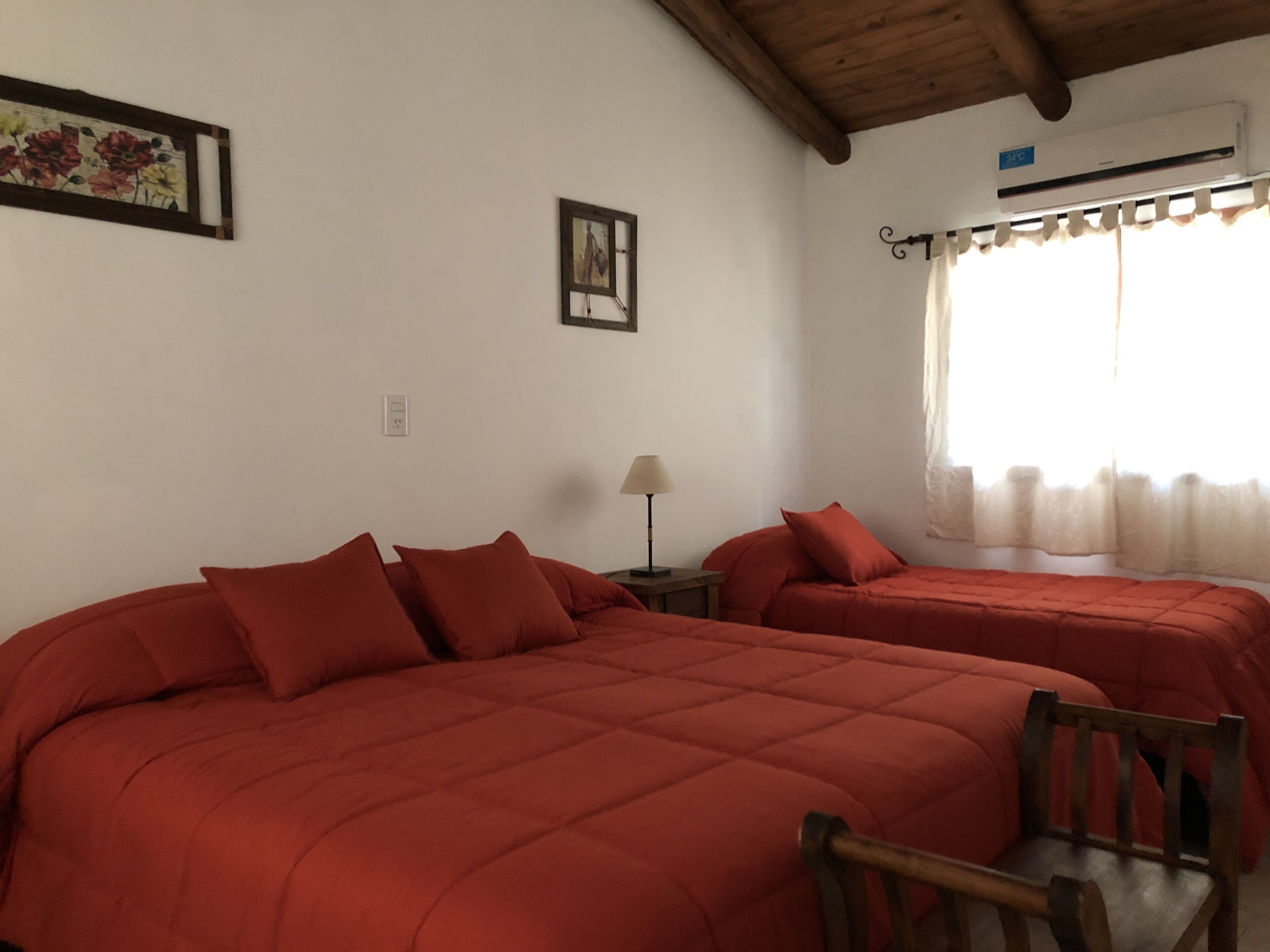 Una habitación con dos camas, una silla de madera, una lámpara y un ventanal con cortinas.
