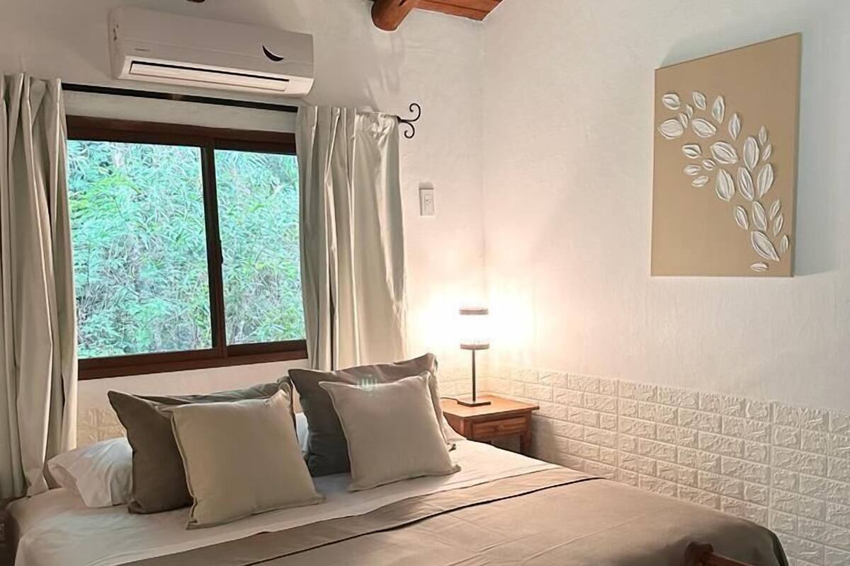 A bedroom with a wooden ceiling, a bed with beige bedding, a wooden bench, a nightstand with a lamp, and a window with curtains.