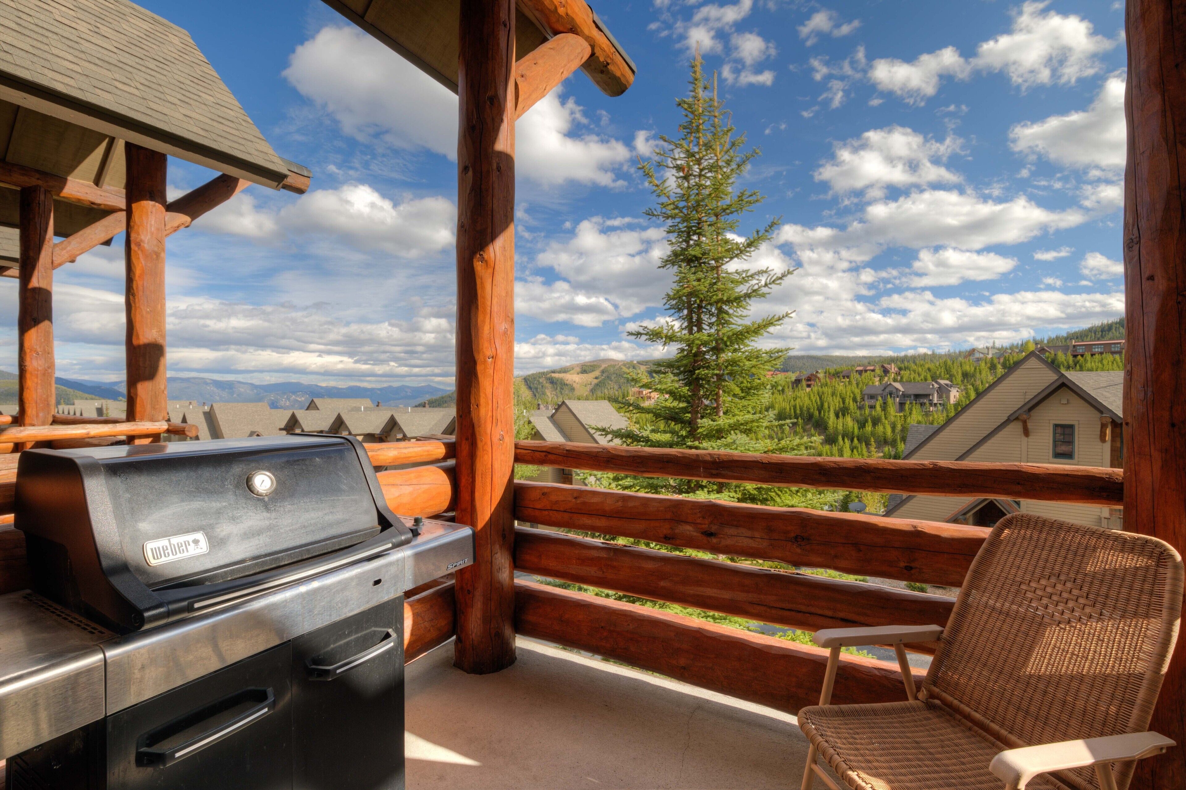 A wooden deck with a grill, a wicker chair, and a view of a forest and houses.