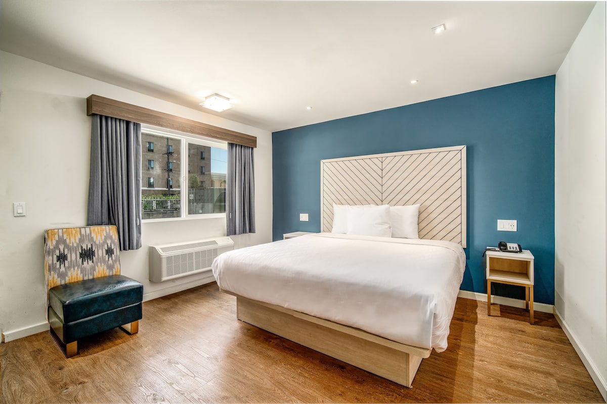 Una habitación de hotel moderna con una cama grande, una silla, una mesita de noche y un ventanal con cortinas.