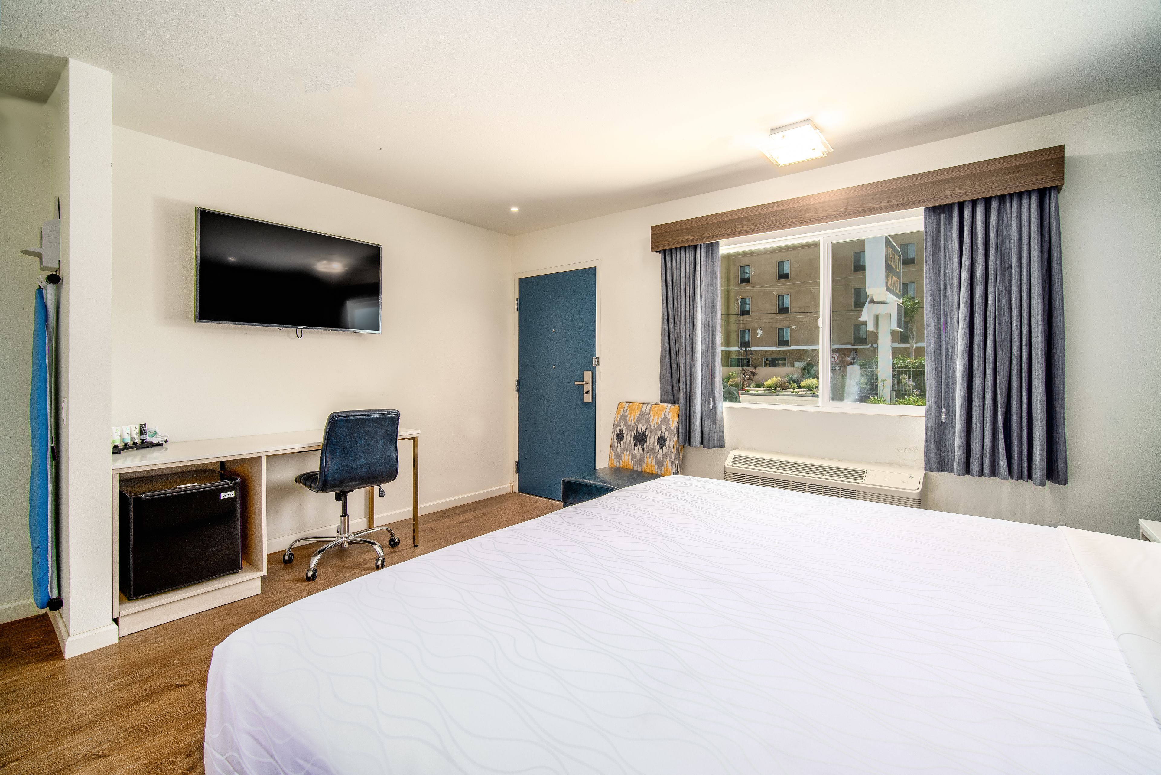 A hotel room with a large bed, a desk with a chair, a flat-screen TV mounted on the wall, and a window with curtains.