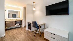 Una habitación de hotel moderna con suelo de madera, un escritorio blanco con silla, un espejo grande, un televisor montado en la pared y un mueble pequeño.