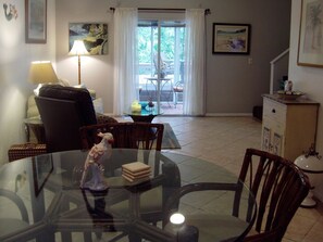A dining area with a glass table, chairs, and a decorative bird statue. There is a lamp, a painting, and a view of a balcony with outdoor furniture.