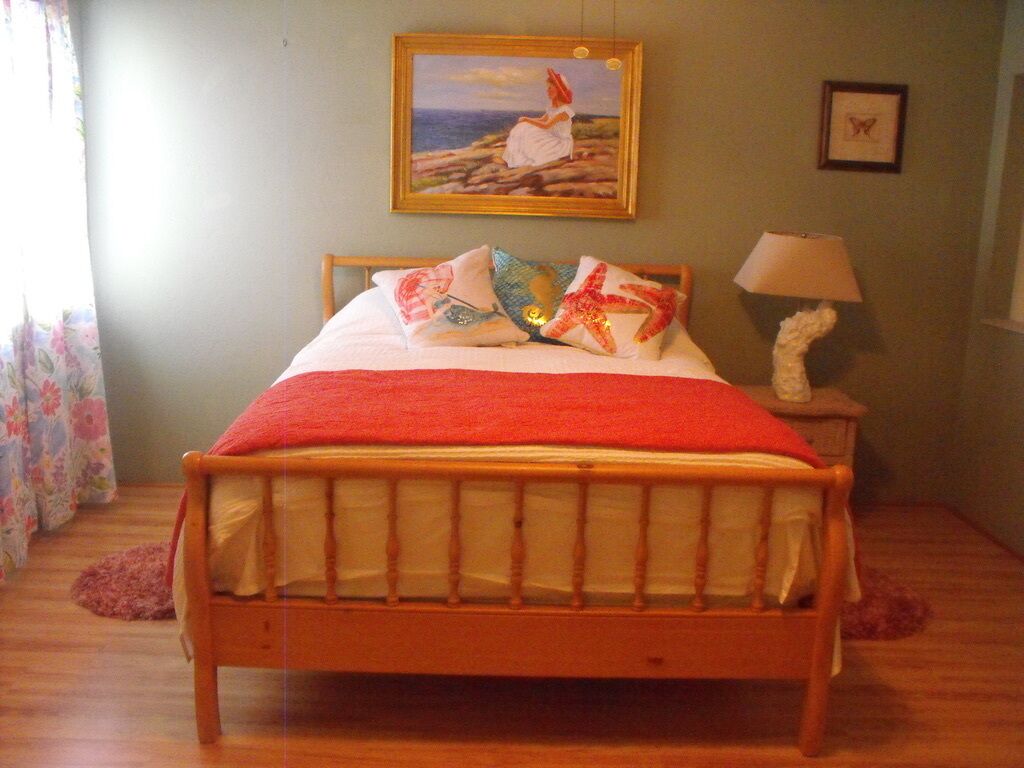 A wooden bed with a red blanket, pillows, and a framed painting of a woman by the sea.