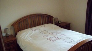 A single bed with a wooden headboard and a white bedspread with geometric patterns. There is a small bedside table with a lamp and an alarm clock on it. - Clougha View (Lancaster)