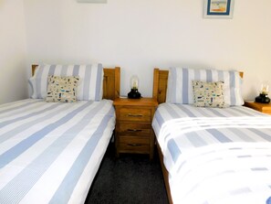 Ein Doppelzimmer mit zwei Betten, jedes mit gestreifter Bettwäsche und Kissen im Seestil. Zwischen den Betten befindet sich ein Nachttisch mit einer Lampe.