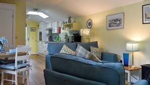 Een woonkamer met een bank, een eettafel met stoelen, een keuken op de achtergrond en een lamp op een bijzettafeltje.