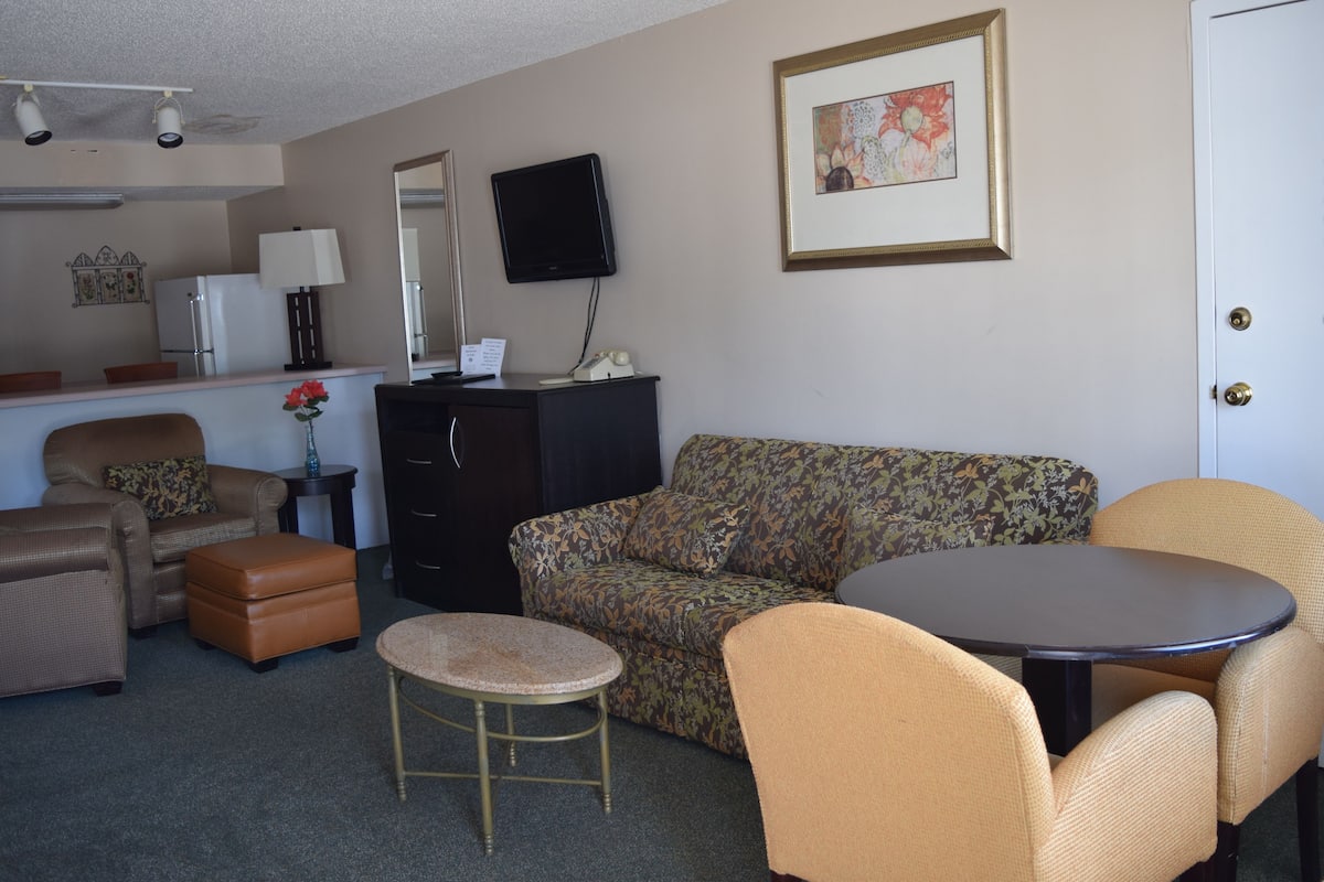 A furnished living room with a sofa, armchairs, a small round table, a coffee table, a TV, a refrigerator, and a cabinet.
