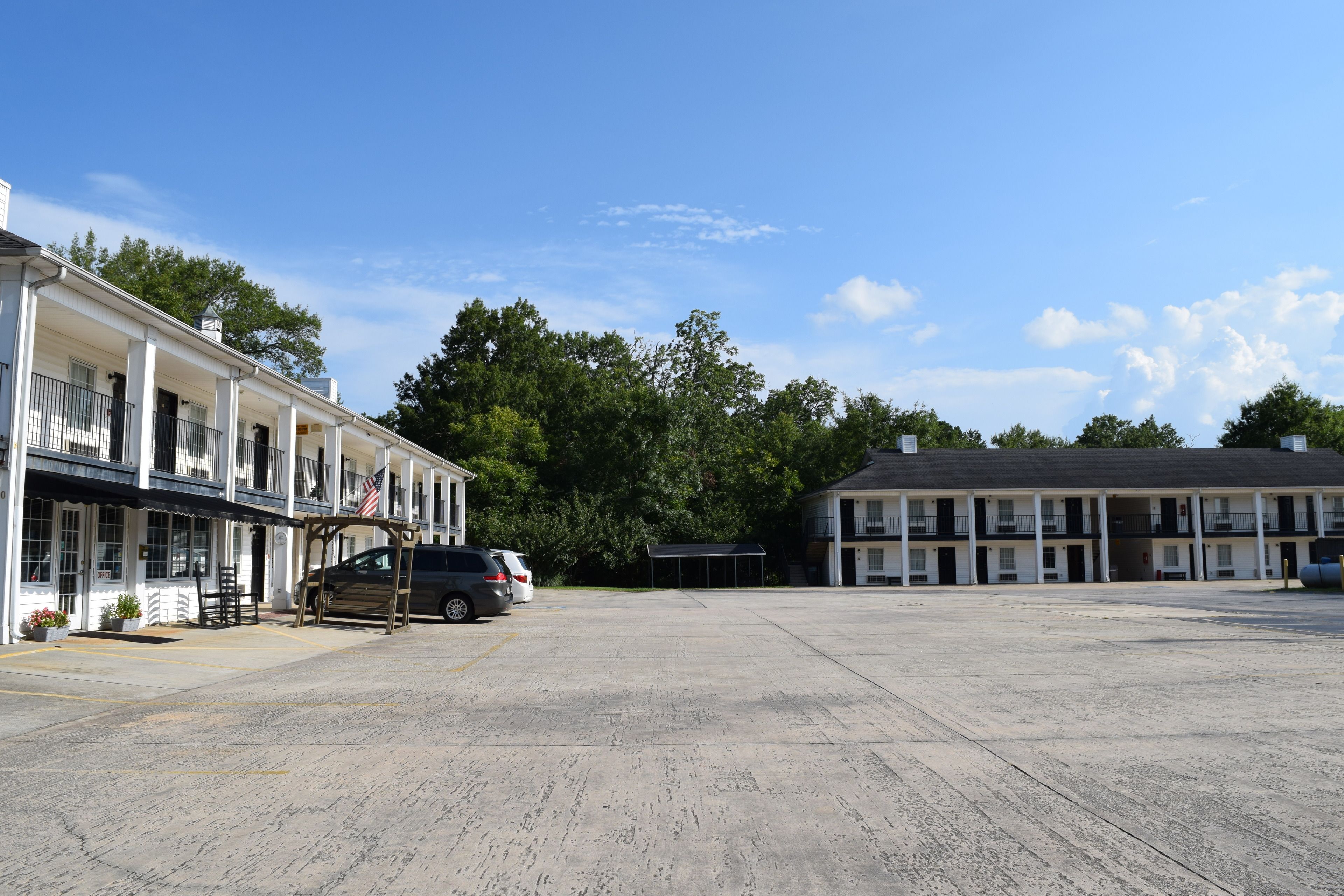Un parking bordé d’une rangée de bâtiments, dont un motel doté d’un espace de stationnement couvert et un bâtiment indépendant avec un porche.
